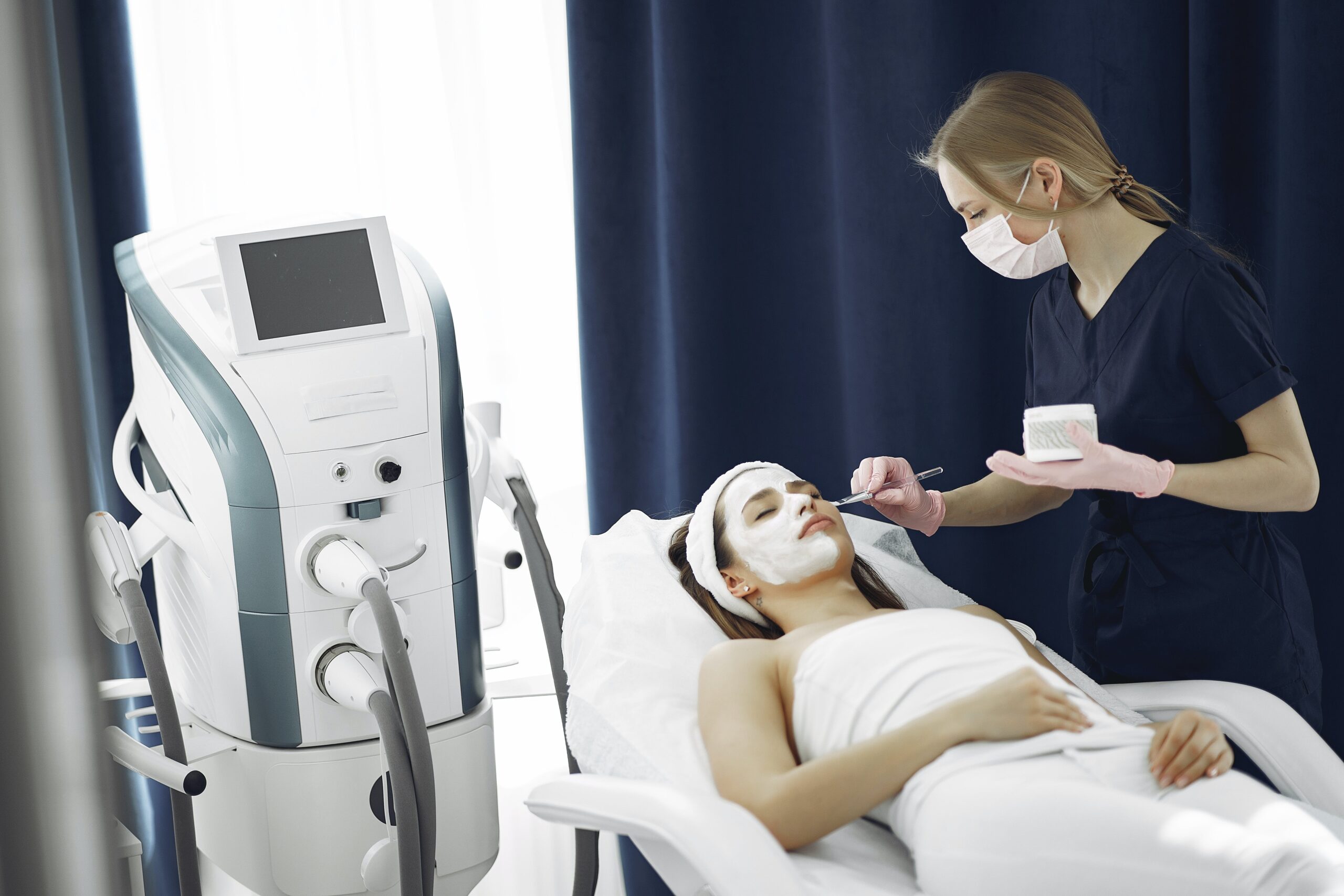 Woman in skin clinic receiving a facial skin treatment from another lady. There are also many skin treatment devices in the room.