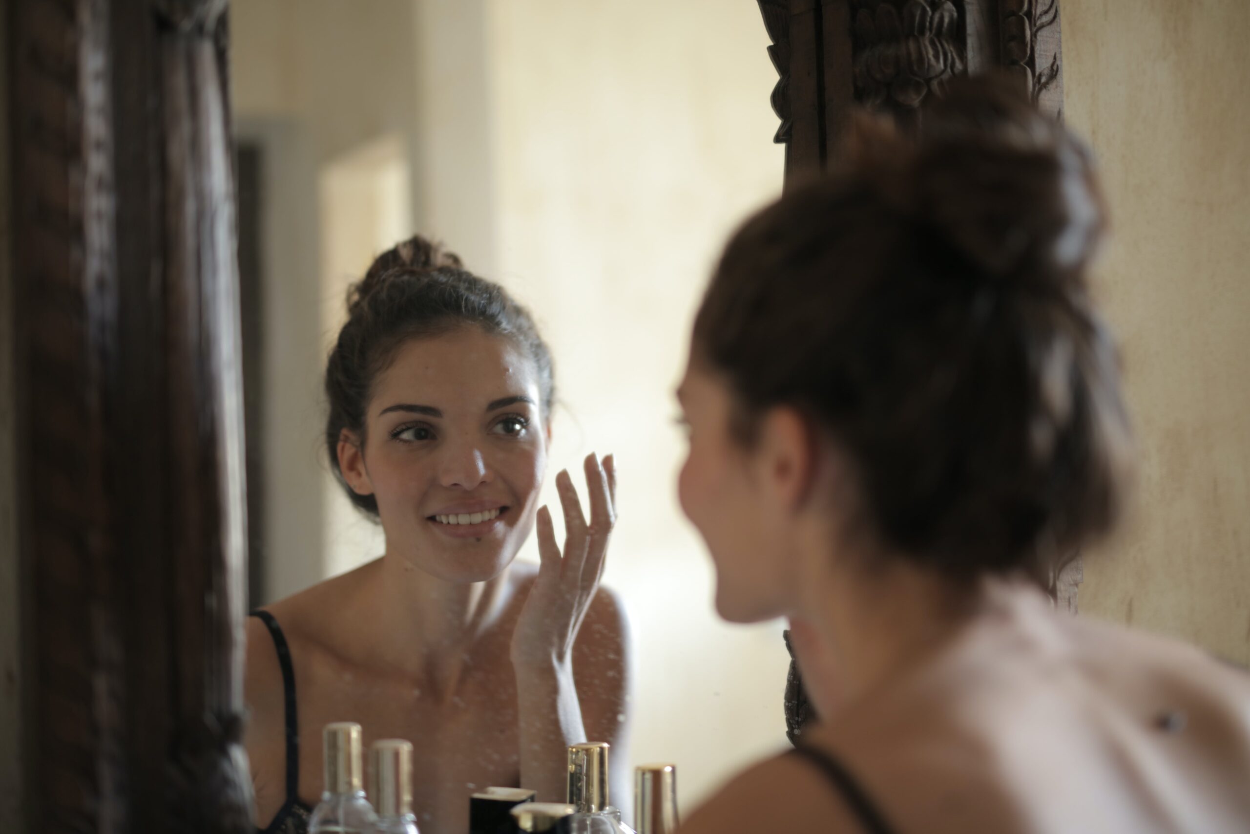 Woman looking in the mirror examining the results of skin cycling on her face. She is happy examining her smooth skin.